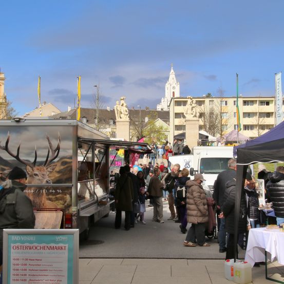 Menschenmengen besuchen einen lokalen Markt. Es gibt Stände und Essenswagen, ein Reh auf einem LKW. Im Hintergrund sind Gebäude und eine Kirche.