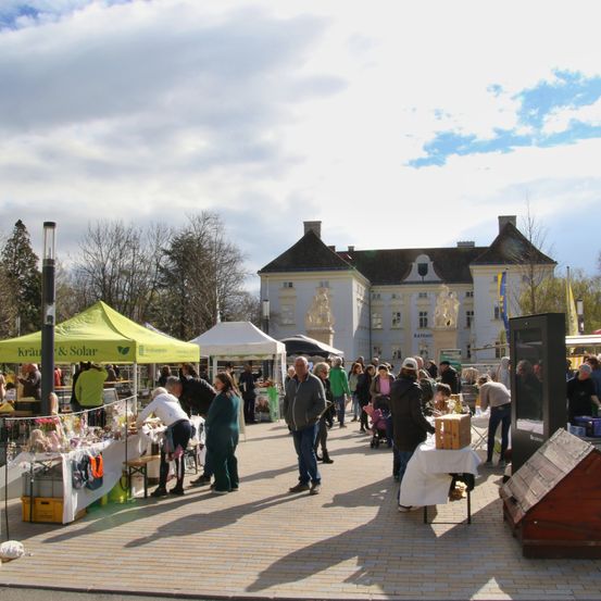 Ein sonniger Outdoor-Markt mit Zelten und Ständen. Menschen stöbern nach Waren und unterhalten sich. Ein großes weißes Gebäude ist im Hintergrund.