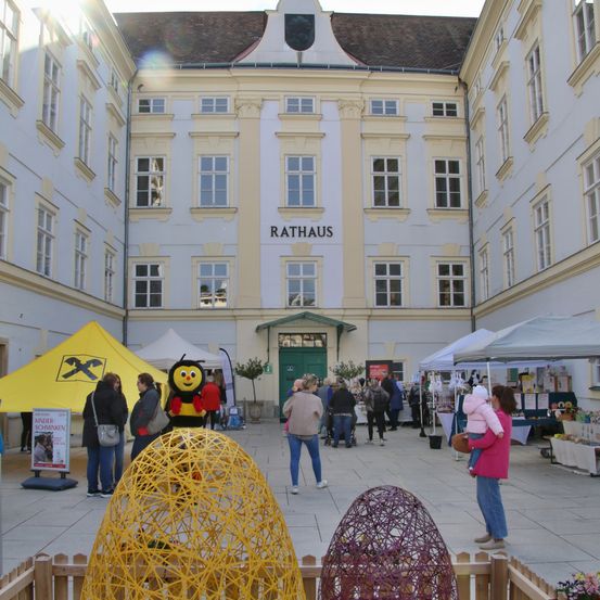 Ein Außenbereich vor einem Gebäude namens Rathaus mit Menschen und dekorativen Objekten wie großen gewebten Eiern. Eine Person in einer Bienenkostüm steht unter ihnen.