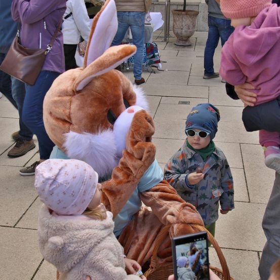 Eine als Hase verkleidete Person interagiert mit zwei Kindern, eines davon hält eine Kamera, in einem öffentlichen Raum. Andere Menschen stehen umher, darunter eine Frau mit einem Baby.
