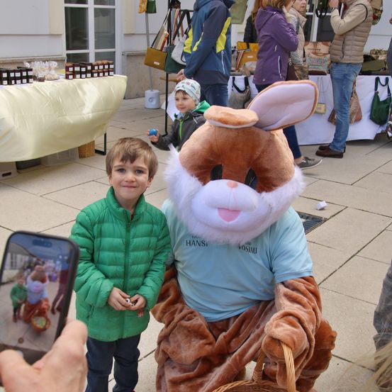Ein Junge in einer grünen Jacke steht neben einer Hasenmaskottchen. Hinter ihnen sind Menschen auf einem Markt unterwegs. Eine Person macht ein Foto mit einem Handy.