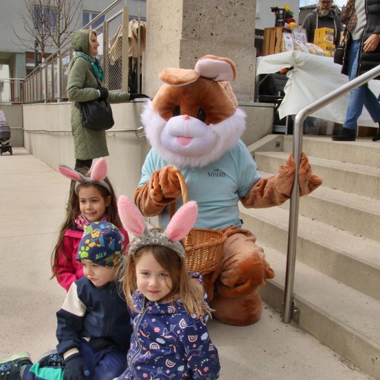 Eine Person in einem Hasenkostüm posiert mit drei Kindern auf einer Betonfläche. Eine Frau geht hinter ihnen auf der Treppe. Ein Kinderwagen und ein Tisch mit Gegenständen sind in der Nähe.
