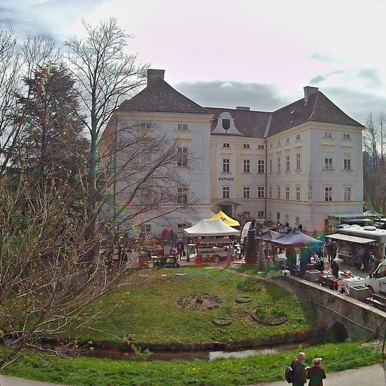 Ein Außenmarkt vor einem großen weißen Gebäude mit vielen Fenstern und einem braunen Dach. Ein Rasenbereich mit einem kleinen Bach und vorbeigehenden Menschen.