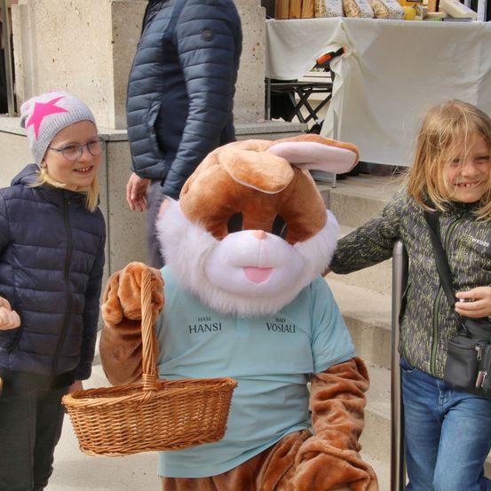 Drei Kinder stehen draußen mit einer Person in einer Hasenkostümmaske, die einen Korb hält. Die Kinder tragen Jacken und eines trägt einen Hut mit einem Stern. Ein Tisch mit Essen steht hinter ihnen.