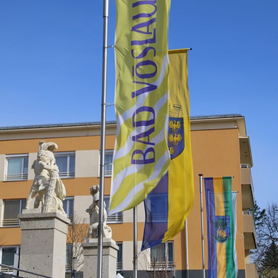 Eine gelbe Fahne mit dem Text 'BAD VOSLAUE' hängt vor einem Gebäude. Die Fahne hat ein weißes und lila Design. Es gibt auch andere Fahnen mit verschiedenen Farben und Designs.