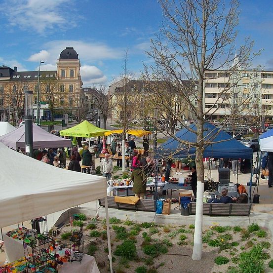 Ein Outdoor-Markt mit verschiedenen Ständen unter farbenfrohen Zelten. Menschen sind um Tische versammelt, die Gegenstände präsentieren. Gebäude und ein Turm sind im Hintergrund zu sehen.