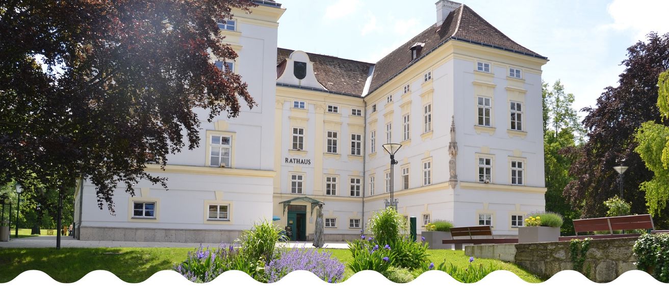 Ein großes weißes Gebäude mit braunem Dach und mehreren Fenstern, auf denen Rathaus steht. Vor dem Gebäude steht eine Statue. Im Vordergrund befinden sich Pflanzen und Blumen.