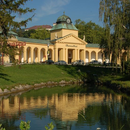 Ein großes gelbes Gebäude mit einem Uhrturm, genannt Thermalbad, steht an einem Teich mit Reflexionen. Bäume und Autos sind in der Nähe.