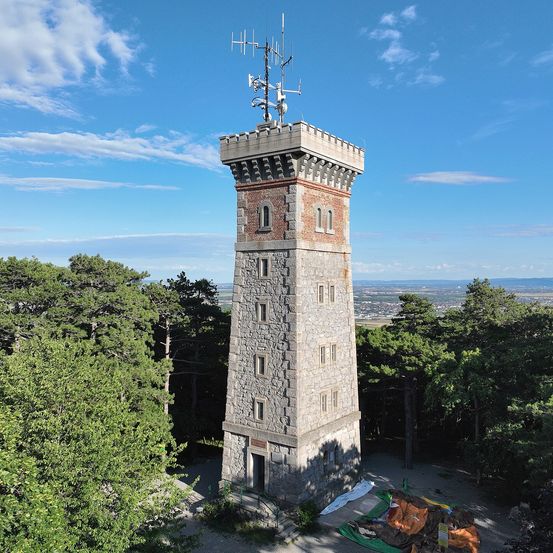 Ein hoher Steinturm mit Antennen steht auf einem Hügel, umgeben von Bäumen unter einem klaren blauen Himmel.