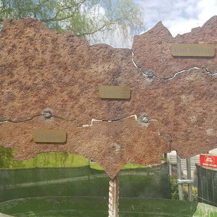 Eine verrostete Metalltafel mit Namen, im Freien aufgestellt. Sie zeigt deutliche Abnutzungserscheinungen. Im Hintergrund befinden sich grüne Pflanzen und ein Gebäude.