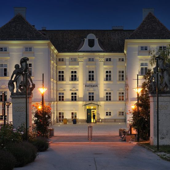 In der Nacht ist die Vorderseite des Rathauses beleuchtet, mit Statuen und Pflanzen auf beiden Seiten und einer Person in der Tür.