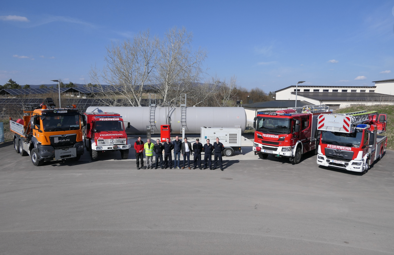 Eine Gruppe Feuerwehrleute steht vor Feuerwehrautos und einem großen Lagertank. Die Umgebung ist im Freien mit einem klaren blauen Himmel.