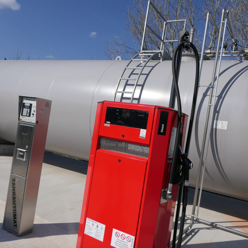 Ein roter Kraftstoffspender steht neben einem großen silbernen Tank. Der Spender ist mit einer Tastatur ausgestattet und hat einen Schlauch angeschlossen. Eine Leiter ist am Tank befestigt.
