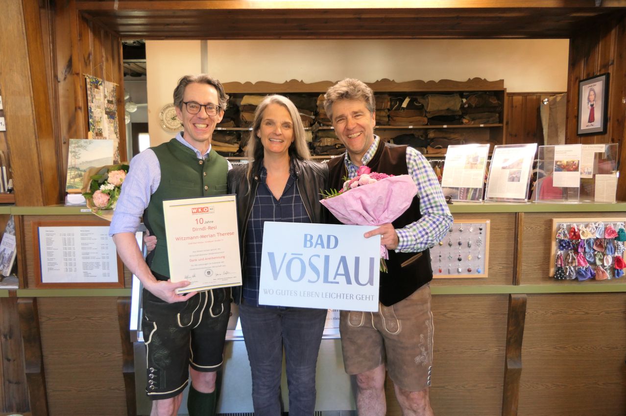 Drei Personen stehen lächelnd eng beieinander in einem Geschäft. Ein Mann auf der linken Seite hält ein Zertifikat. Die Frau in der Mitte hält einen Blumenstrauß. Der Mann auf der rechten Seite hält ein Schild mit der Aufschrift 'Bad Vöslau.'