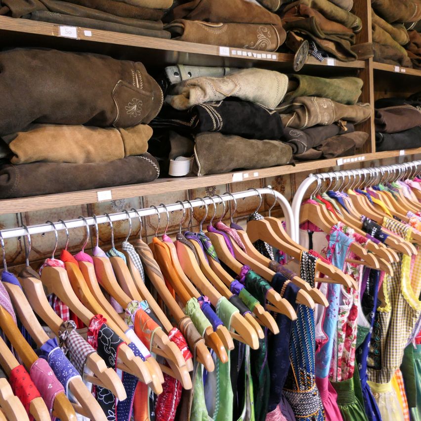 Ein Bekleidungsgeschäft zeigt eine Vielzahl von Kleidungsstücken auf Holzregalen und Metallbügeln. Verschiedene Farben und Arten von Kleidung sind sichtbar, einschließlich Lederstiefel.