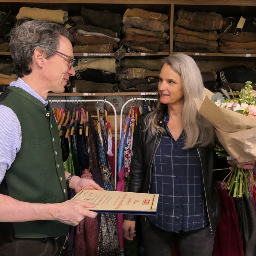 Ein Mann mit Brille und grüner Weste überreicht einer lächelnden Frau, die einen Blumenstrauß hält, ein Zertifikat in einem Bekleidungsgeschäft.