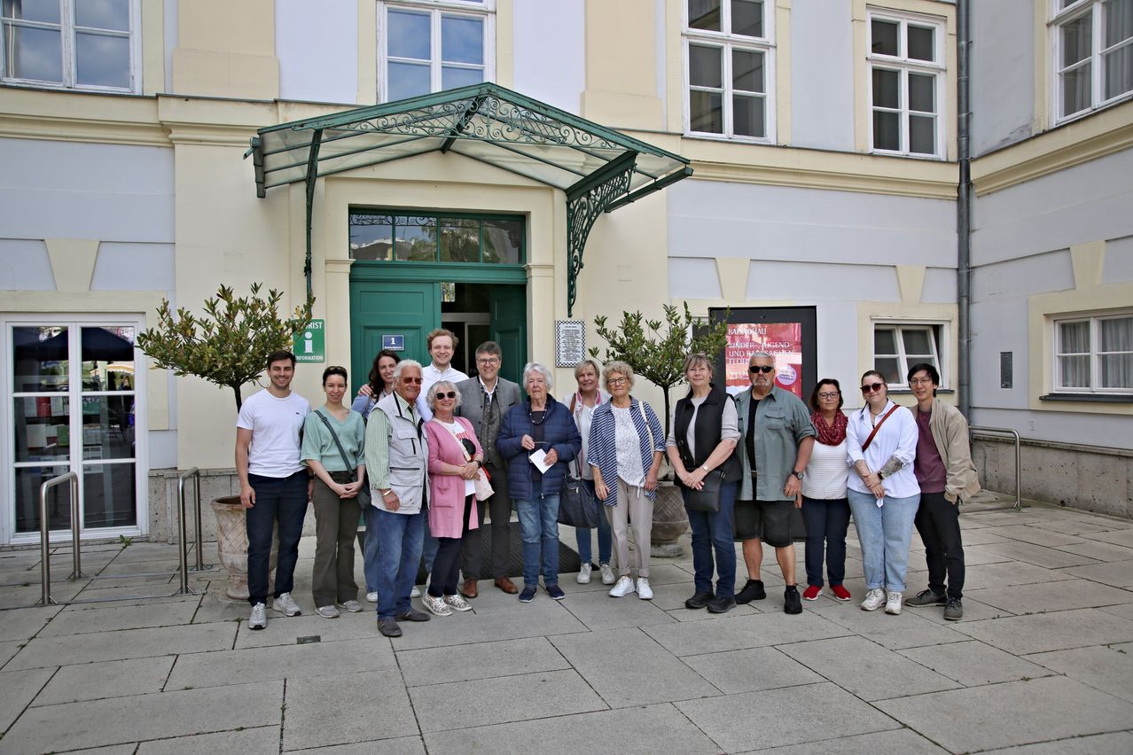 Eine Gruppe von Menschen steht vor einem Gebäude, lächelt für ein Foto. Sie tragen Freizeitkleidung und stehen in einer Reihe. Das Gebäude hat einen grünen Eingang und mehrere Fenster.