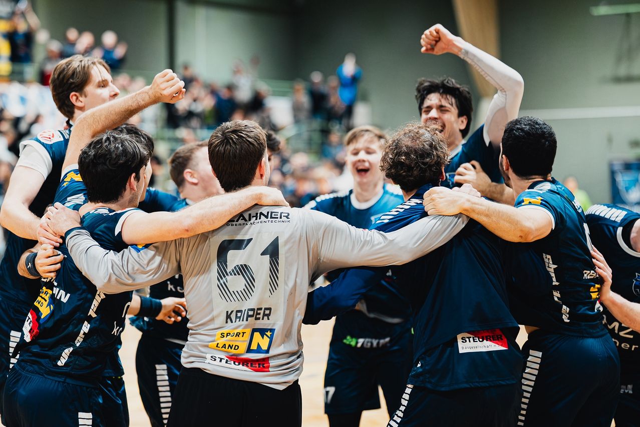 Eine Gruppe Handballspieler feiert in einer voll besetzten Arena, ein Spieler trägt ein graues Trikot mit der Nummer 61.