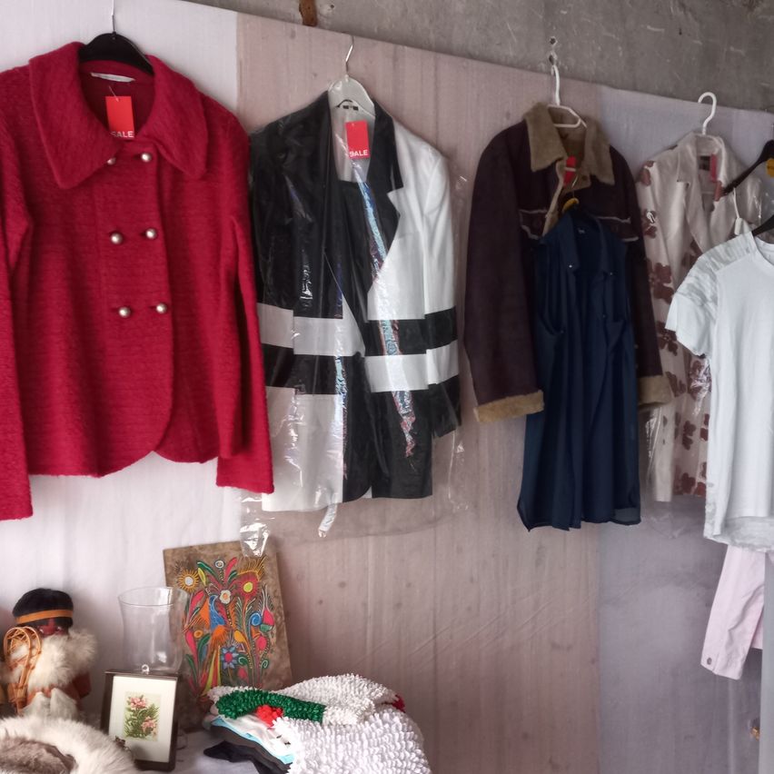 Eine Wand mit verschiedenen Kleidungsstücken, darunter ein roter Mantel, ein schwarz-weißes Jackett und ein braunes, mit Fell gefüttertes Jackett. Darunter sind einige gefaltete Kleidungsstücke und ein Bilderrahmen.