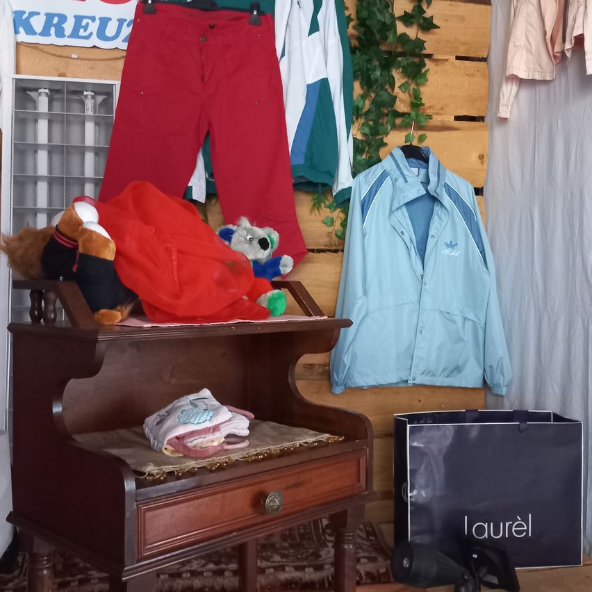 Ein hölzerner Schrank mit verschiedenen Kleidungsstücken und Spielzeug oben drauf. Eine blaue Jacke hängt an der Wand und ein schwarzer Beutel mit dem Wort 'Laurel' steht daneben.