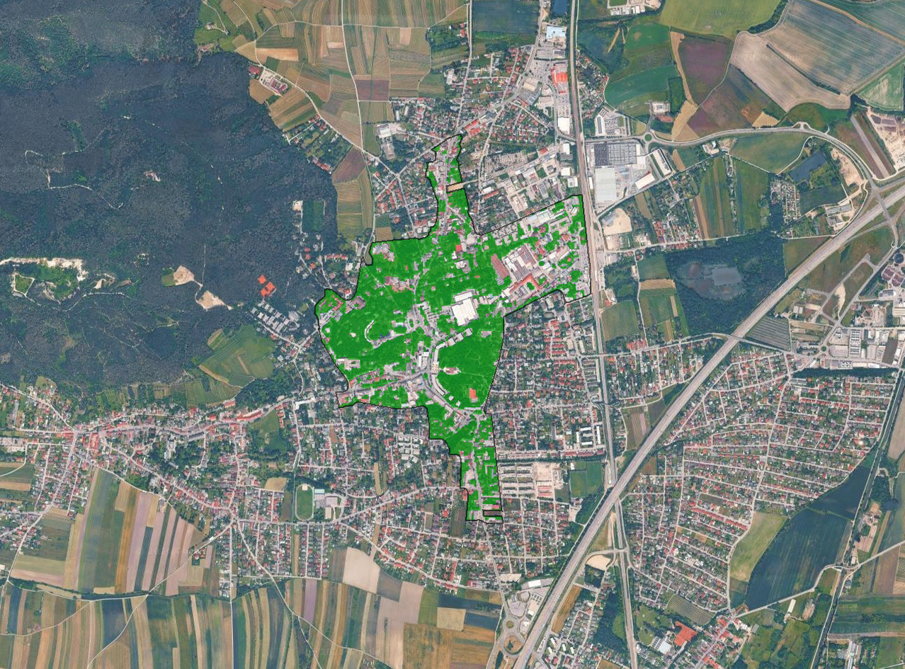 Ein Luftbild einer Stadt mit verschiedenen grünen Flächen. Der zentrale Bereich ist dicht besiedelt mit Gebäuden und hat einen großen grünen Park. Um die Stadt herum befinden sich landwirtschaftliche Felder und ein Gewässer.