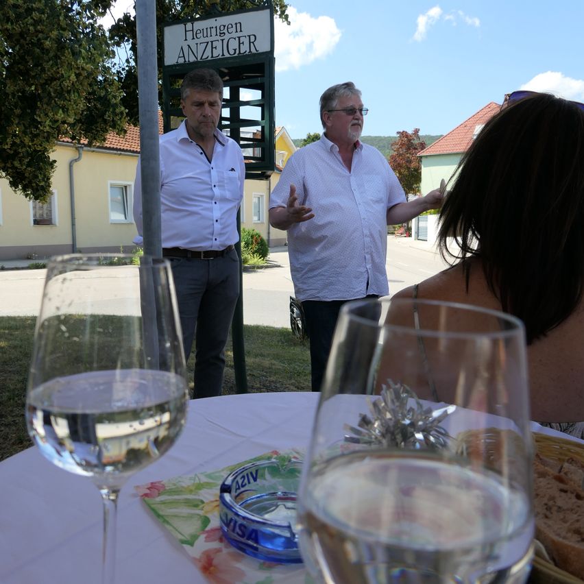 Zwei Männer in weißen Hemden und mit Brille stehen vor einem Tisch mit Weingläsern und Brot. Eine Frau hört ihnen zu, während sie sitzt. Das Schild heißt Heurigen Anzeiger.