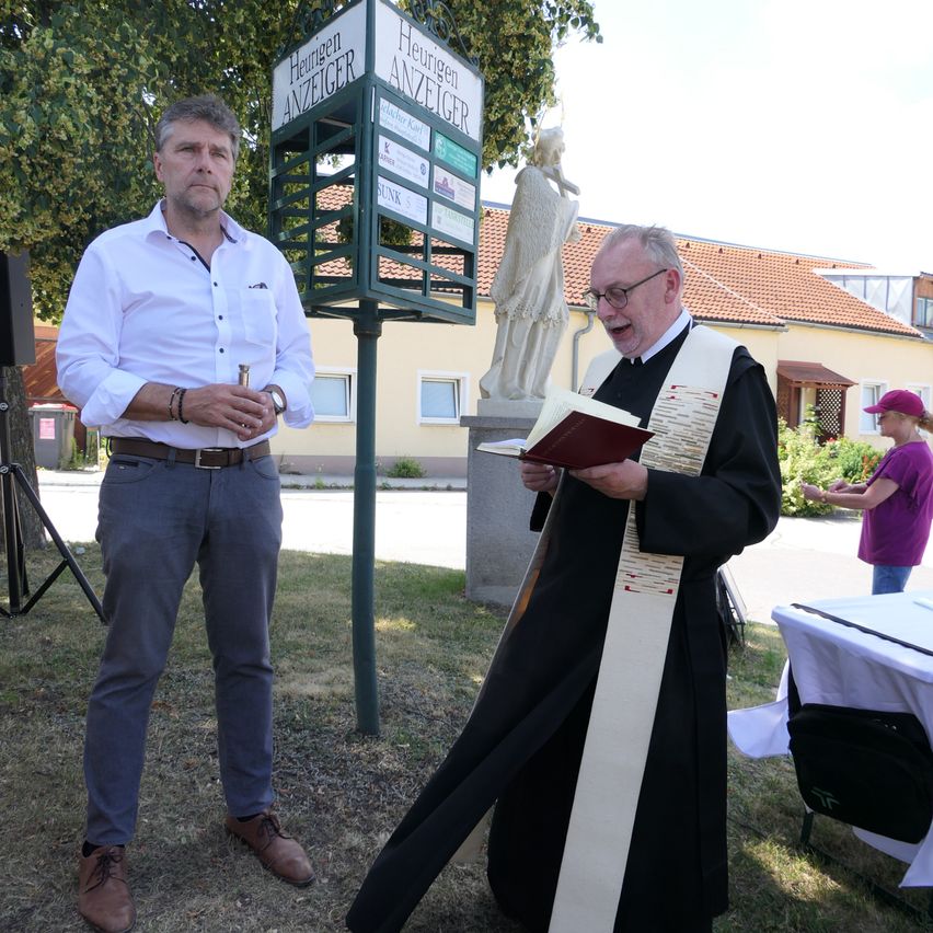 Ein Mann in einem weißen Hemd steht neben einer Tafel, während ein Mann in einer schwarzen Robe ein Buch liest. Hinter ihnen steht eine Statue und ein Gebäude.