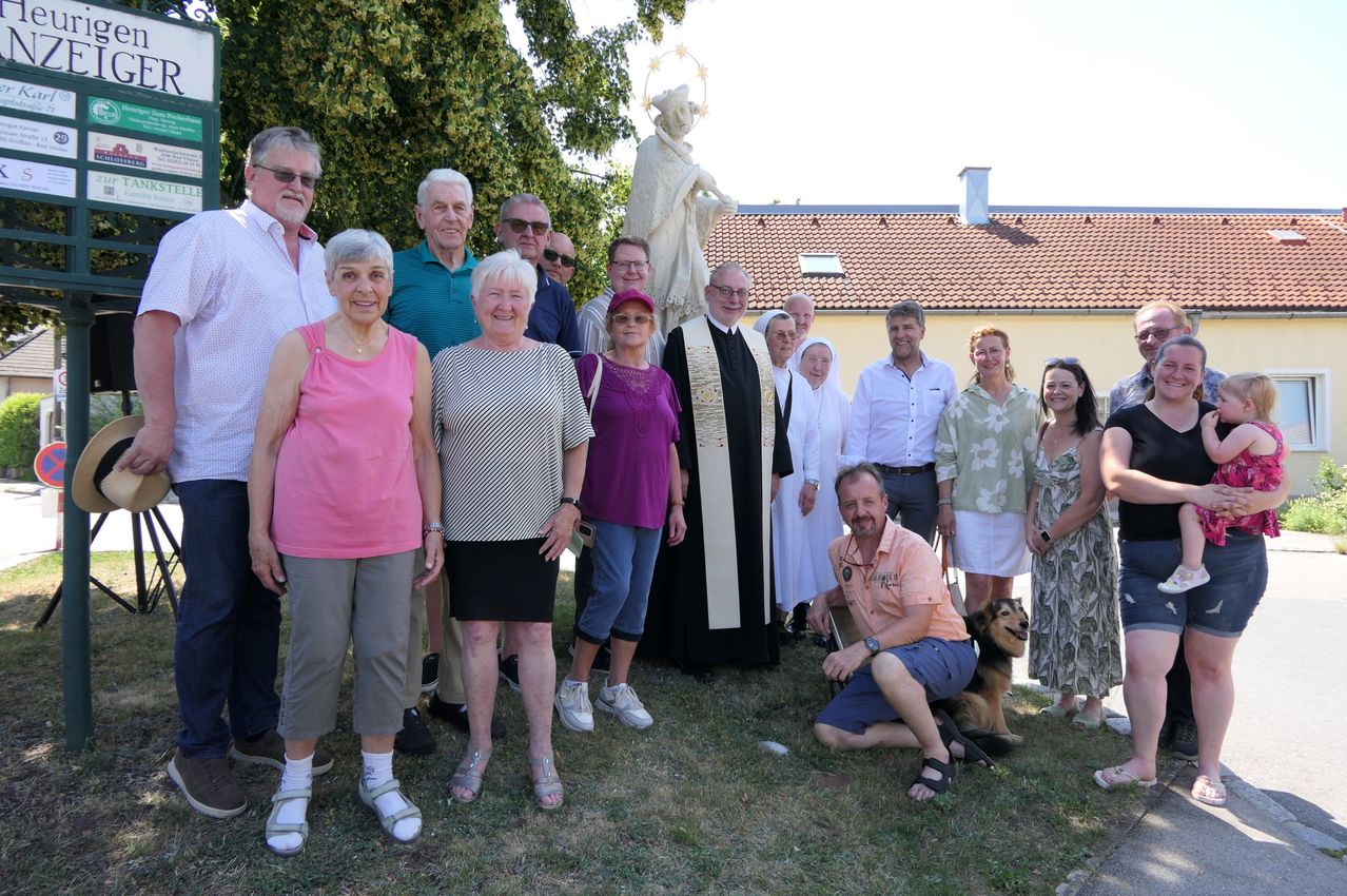 Eine Gruppe von Menschen, einige mit Brillen und Hüten, posiert für ein Foto vor einer Statue und einem Gebäude. Ein Mann kniet mit einem Hund, und eine Frau steht mit einer Handtasche.