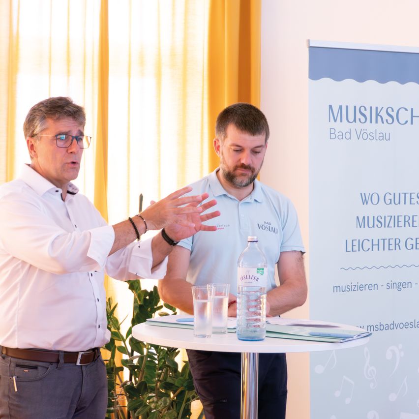 Zwei Männer stehen vor einem Tisch mit Wassergläsern und einer Flasche, einer erklärt dem anderen etwas. Dahinter befindet sich ein Plakat mit den Worten 'Musikschau Bad Vöslau' und einem Musiknotensymbol.