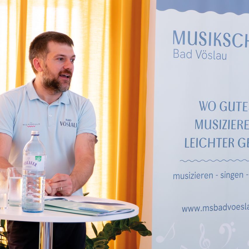 Ein Mann steht vor einem Tisch mit einer Flasche Wasser und einem Glas. Er trägt ein weißes Poloshirt. Hinter ihm befindet sich ein Banner mit der Aufschrift 'Musikschule Bad Vöslau' und ein Topf mit einer Pflanze. Das Bild hat einen Wasserzeichen unten links.