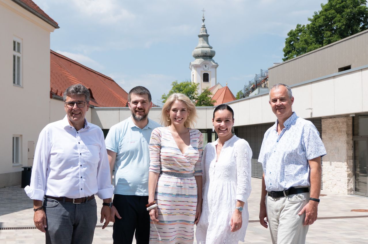 Fünf Menschen stehen vor einem Gebäude. Drei Männer und zwei Frauen. Hinter ihnen steht ein Turm.