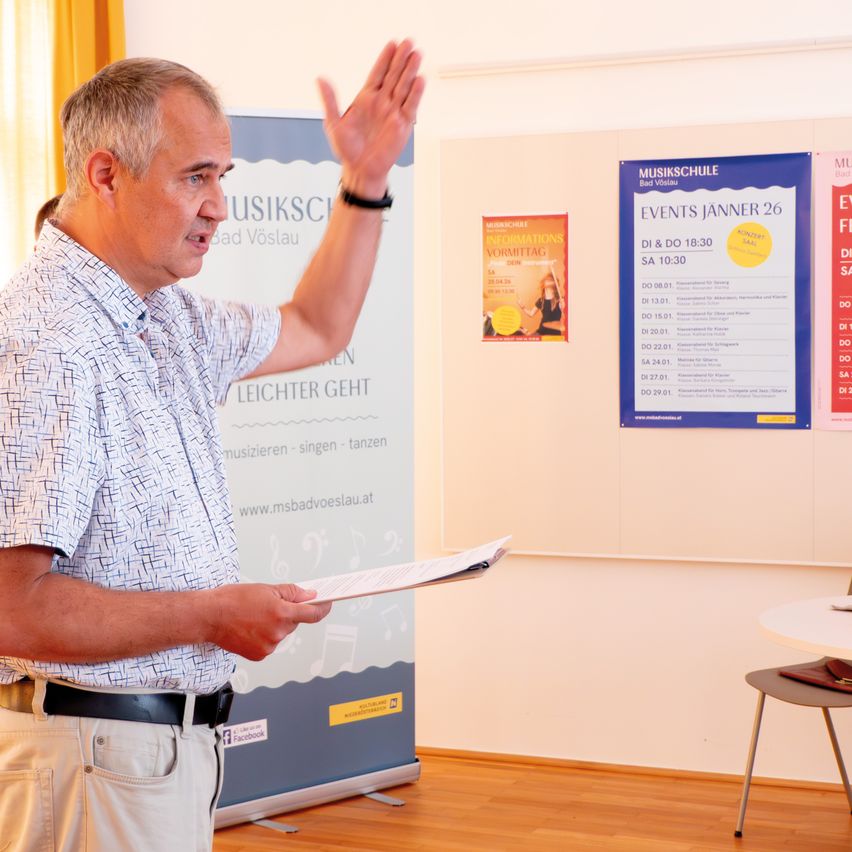 Ein Mann steht in einem Raum, hält ein Papier in der Hand und spricht mit einer Handgeste. Hinter ihm ist eine weiße Wand mit einem Plakat befestigt.