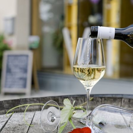 Eine Flasche Weißwein wird in ein Weinglas auf einem Holzfass gegossen, mit einer roten Rose und einer weißen Blume in der Nähe, mit einem verschwommenen Gebäude und einer Tafel im Hintergrund.