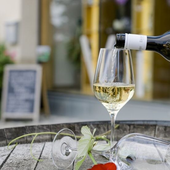 Eine Flasche Weißwein wird in ein Weinglas auf einem Holzfass gegossen, mit einer roten Rose und einer weißen Blume in der Nähe, mit einem verschwommenen Gebäude und einer Tafel im Hintergrund.