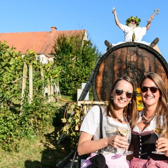 Zwei Frauen halten Weinglas vor einem Fass. Ein Mann in einem Blattkranz sitzt auf dem Fass. Im Hintergrund steht ein Haus und ein Garten mit Pflanzen und einem Holzzaun.