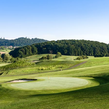 Ein Panoramablick auf einen Golfplatz mit einem üppigen grünen Feld, einem Sandbunker und einer Fahne in der Mitte. Der Platz ist von Bäumen und Hügeln umgeben, mit einem klaren blauen Himmel darüber.