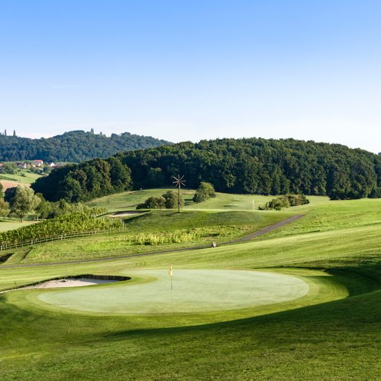 Ein Panoramablick auf einen Golfplatz mit einem üppigen grünen Feld, einem Sandbunker und einer Fahne in der Mitte. Der Platz ist von Bäumen und Hügeln umgeben, mit einem klaren blauen Himmel darüber.
