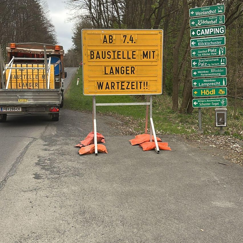 Ein Straßenschild weist auf Baustellen mit längeren Wartezeiten hin. Ein LKW mit einem gelben Schild steht in der Nähe. Straßenschilder weisen auf verschiedene Ziele hin.