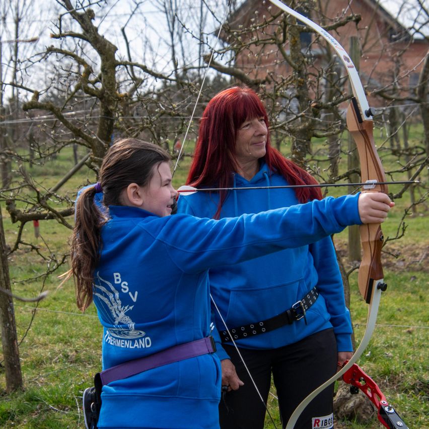 Eine Frau mit roten Haaren übt Bogenschießen, hält einen Bogen und neben ihr steht ein junges Mädchen in Blau. Sie stehen in einem Außenbereich mit Bäumen und einem Haus im Hintergrund.