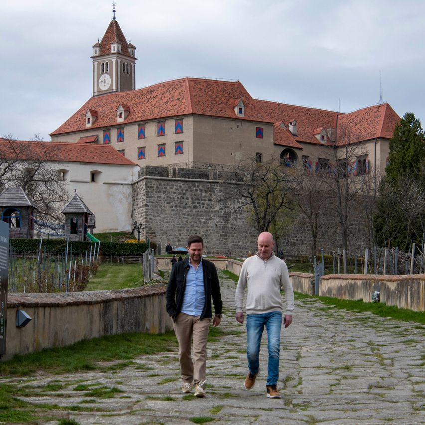 Zwei Männer gehen auf einem Steinpfad vor einem historischen Gebäude mit einem Glockenturm. Das Gebäude hat ein rotes Dach und farbenfrohe Fenster.