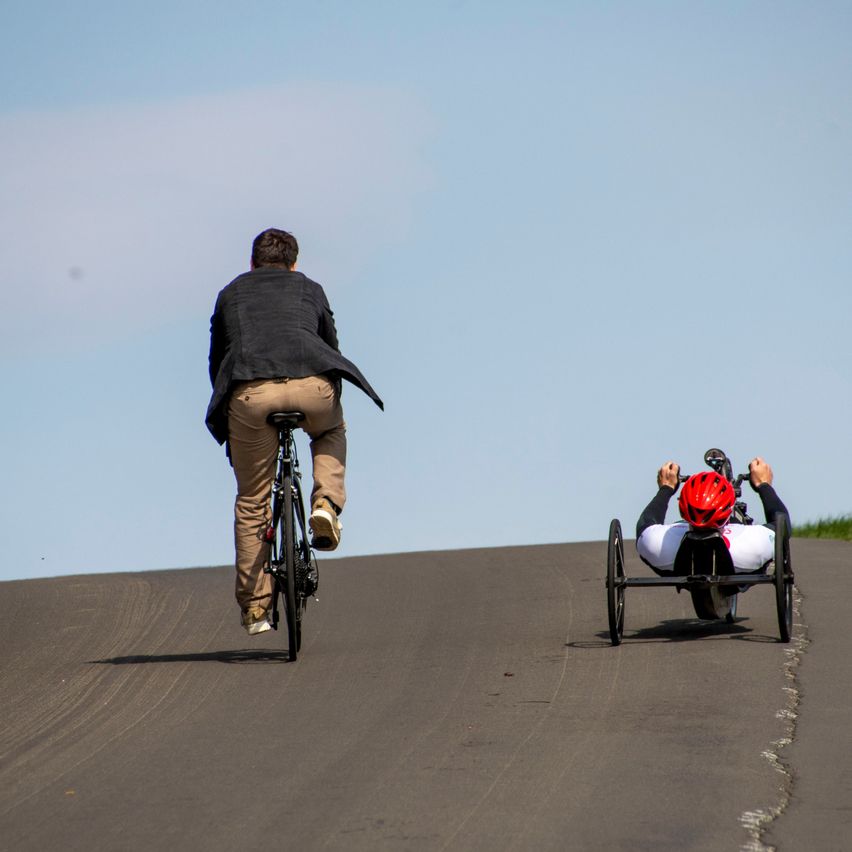 Zwei Radfahrer, einer auf einem Fahrrad und der andere in einem Handfahrrad, fahren auf einer Straße. Der Fahrradfahrer trägt eine schwarze Jacke und beige Hose. Der Handradfahrer trägt ein weißes Shirt, schwarze lange Ärmel und einen roten Helm.
