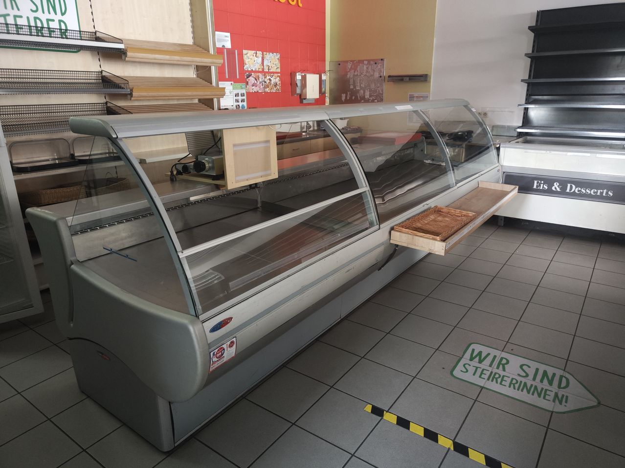 Eine kommerzielle Kühltheke mit einer Glasvitrine und einem hölzernen Tablett im Inneren, in einem Geschäft gelegen. Der Laden hat geflieste Böden und Regale mit verschiedenen Artikeln. Es gibt ein Schild an der Wand mit der Aufschrift 'Eis'.