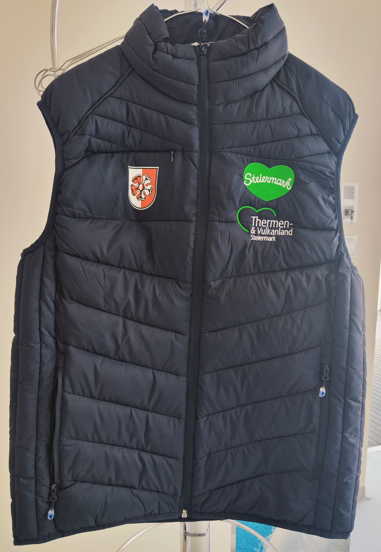 Eine marineblaue Steppweste mit einem roten und weißen Emblem auf der linken Seite und einem grünen herzförmigen Patch mit dem Text 'Stelmark' auf der rechten Seite.