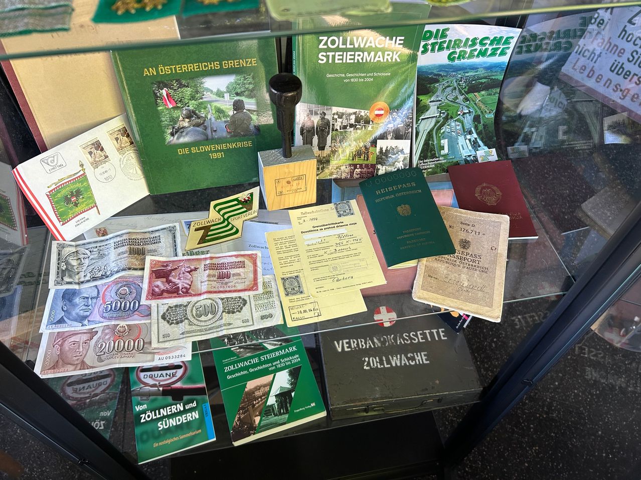 A glass case displays various historical documents, books, stamps, and currency. Notable items include a book titled 'Die Slowenienkrise 1991' and a rubber stamp.