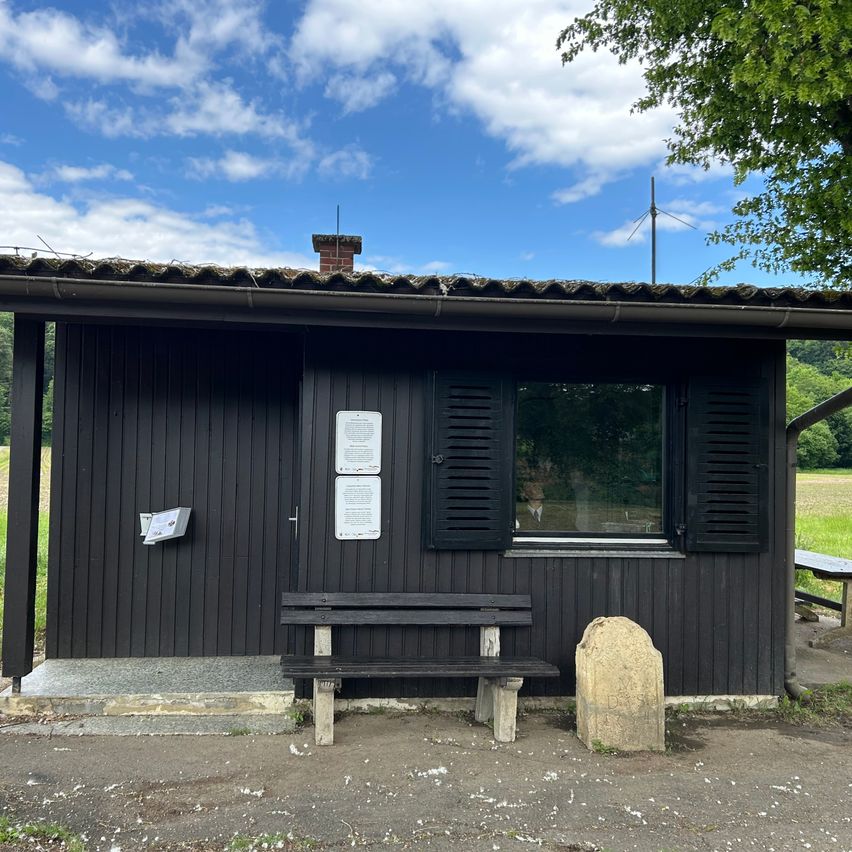 A small black building with a chimney, a window, and two shutters, and a bench in front. There is a sign on the left and a rock on the right.