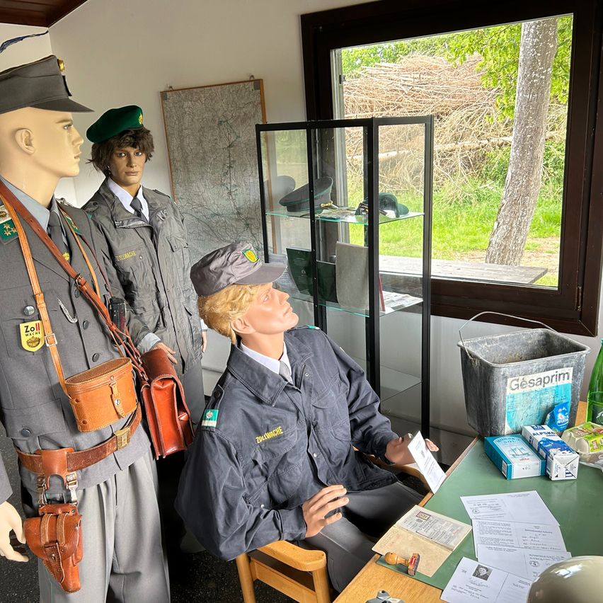 Three mannequins dressed in military uniforms are in a room with a window and a desk. One sits while the other two stand, one holding a rifle. Papers and boxes are on the desk.
