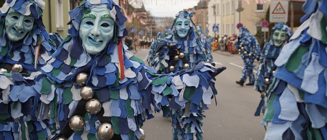 Menschen in blauen und grünen Kostümen mit Glocken gehen bei einer Straßenparade. Die Kostüme sind aufwendig mit Rüschen und Masken. Dahinter sind weitere Teilnehmer zu sehen, und die Straße ist von Gebäuden gesäumt.