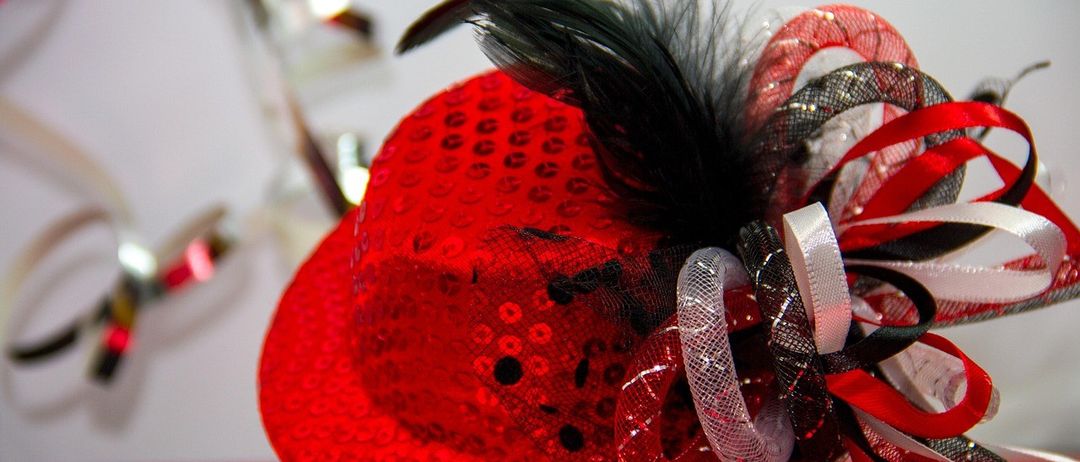 Ein roter Hut mit schwarzen Federn und roten und schwarzen Bändern. Der Hut ist mit schwarzen Punkten verziert und hat eine glänzende Textur.