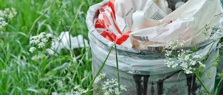 A trash can filled with plastic bags is surrounded by white flowers and green grass. The trash has black stains and is half-buried in the ground.