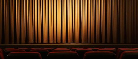 An empty movie theater with rows of red seats under closed curtains and dim lighting.